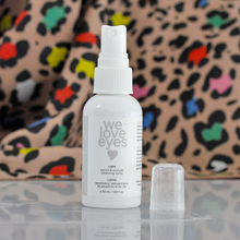 Load image into Gallery viewer, White spray bottle labeled 'we love eyes' on a reflective surface with a colorful leopard print background.
